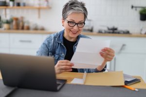 Happy mature woman watching mail bank notice, sitting at kitchen table at home.